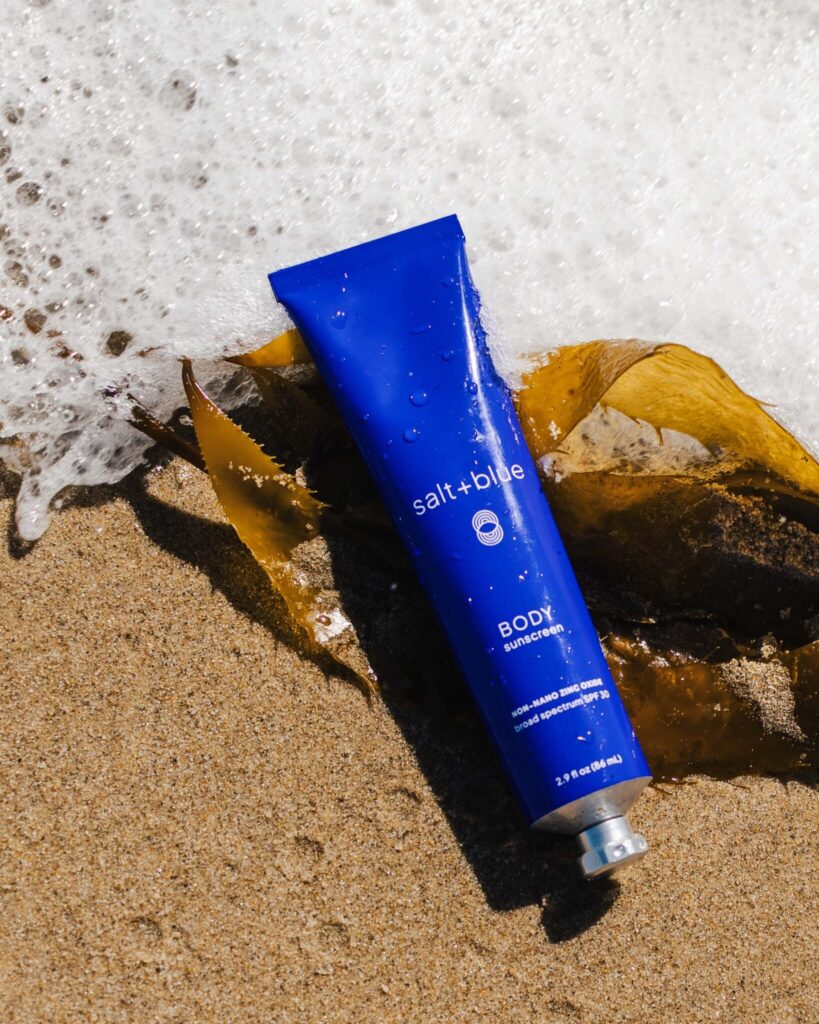 salt+blue BODY Sunscreen against seafoam and kelp on sand